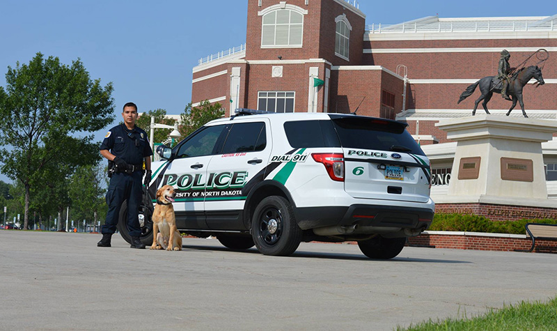 University Police Officer Jose Solis and K-9 officer stand -- and sit -- at the ready near their police vehicle on campus. Solis and Ben are part of a stepped up strategic partnership between between Public Safety and University Information Technology (UIT) to build up UND’s physical and cybersecurity protection systems and enhance public safety on campus. Image courtesy of University Police.