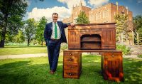 UND’s current President Andy Armacost is pictured with a desk that likely once belonged to the first president of UND, William Blackburn, who served UND from 1884-85. The photo was taken in front of Merrifield Hall and the Eternal Flame. Photo by Sam Melquist/UND Alumni Association & Foundation