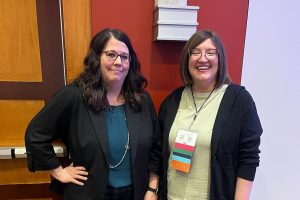 Two women stand in front of colored wall.