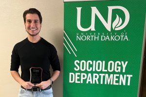 man holds award and stands in front of UND Sociology banner.