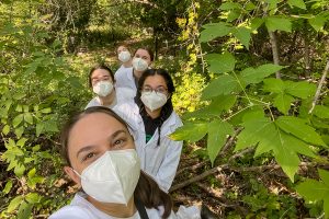 Scientists in white lab coats and masks surrounded by greenery on a trail.