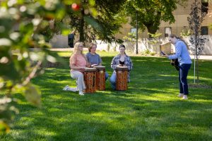 Three students kneeling on grass with drums, while music therapy instructor plays a guitar