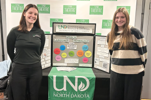 Two CSD students stand along either side of a posterboard presentation about Speech Language Pathology with a UND Theatre backdrop
