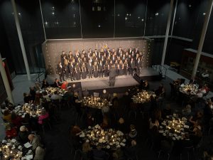 Concert Choir lined on a stage in black uniforms, singing for a crowd of candlelight dinner tables
