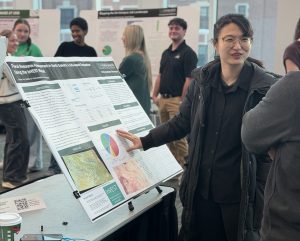 Student with hand on posterboard pointing out research and talking with visitor