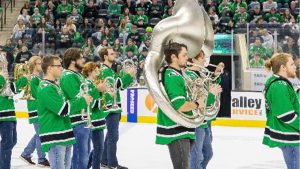 Marching band in hockey jerseys on the ice of Ralph Engelstad Arena, lined up in front of filled stands of hockey fans.