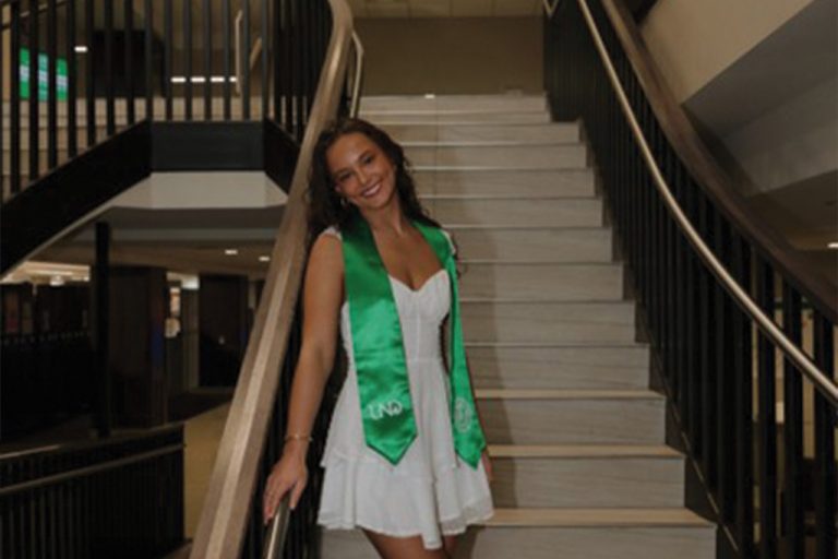 Undergraduate Student Katherine Mary Blackmun standing in white dress and green UND stole on a staircase at Chester Fritz Library