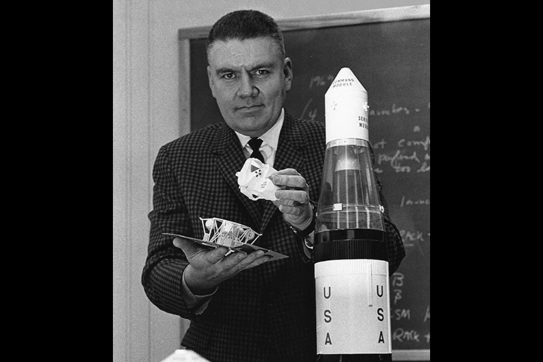 A vintage black and white photograph of a man presenting several spacecraft in front of a chalkboard filled with complex equations.