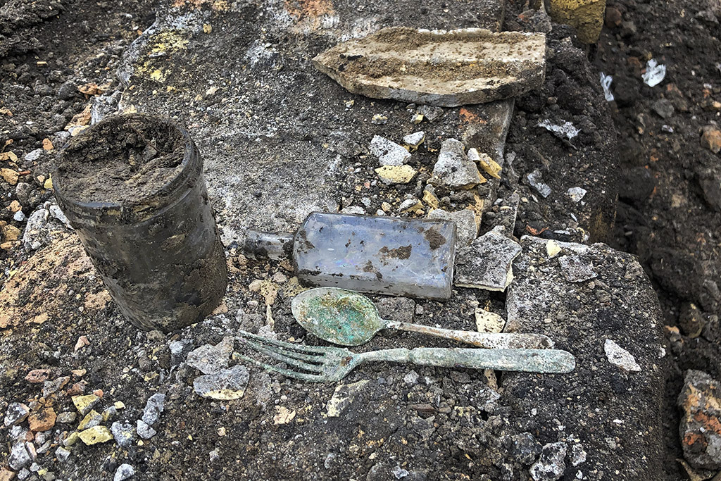 A glass jar, rusted fork, rusted spoon and glass flask, all covered in dirt and debris, sit above an excavation zone