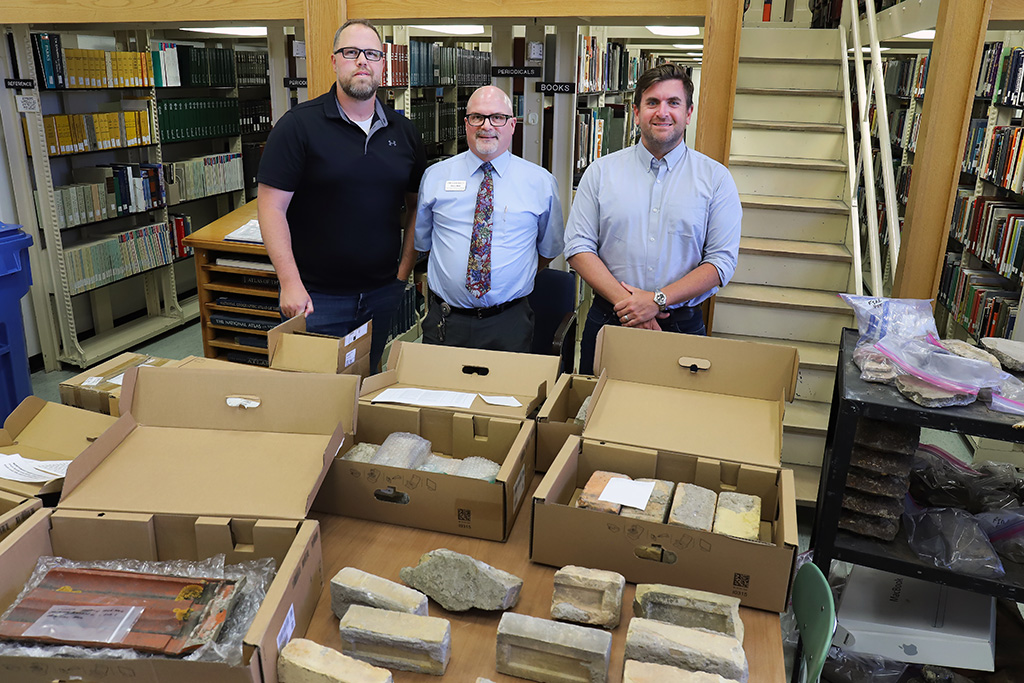 Three UND employees stand behind boxes of brick and other artifacts