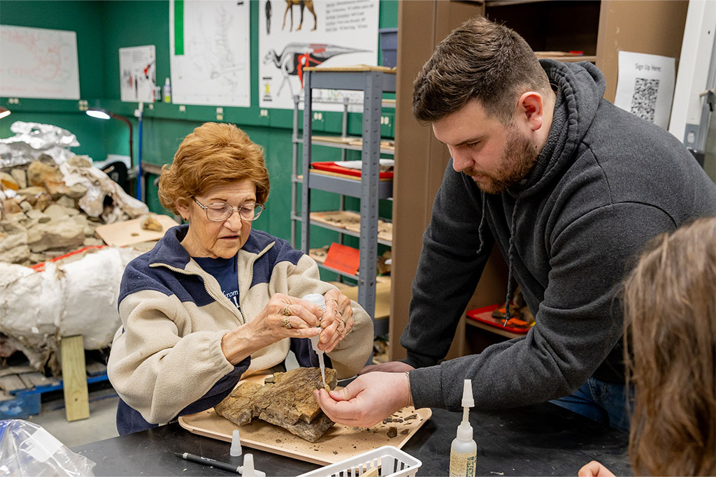 An older woman carefully applies glue to a piece of fossil with the help of a UND graduate student who assists with volunteers assembling fragments of bone.