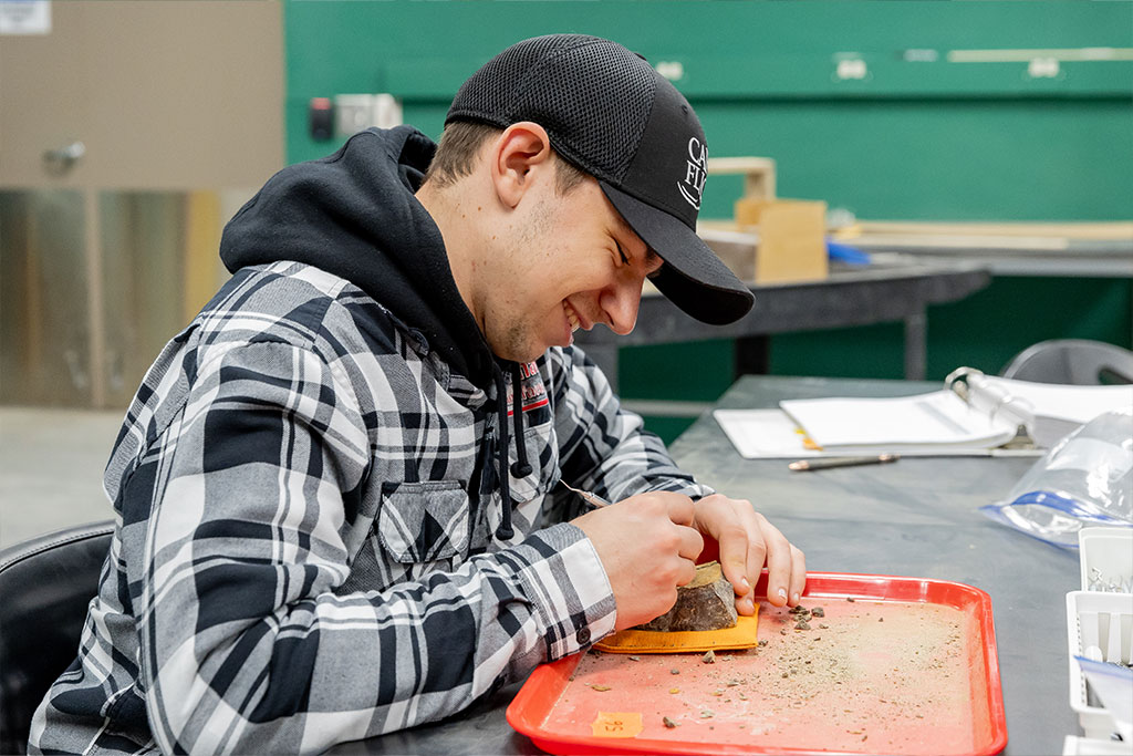 Student smiles as he uses a metal tool to scrape away at rock that surrounds pieces of dinosaur fossils