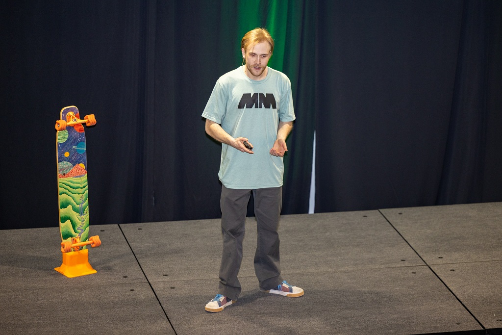 A student talks on a stage. Next to him is an orange skateboard with an art decal on the bottom side on display.