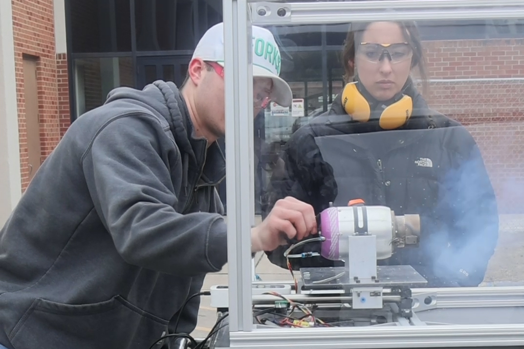 Students work on a rocket engine outdoors as smoke billows out of the engine