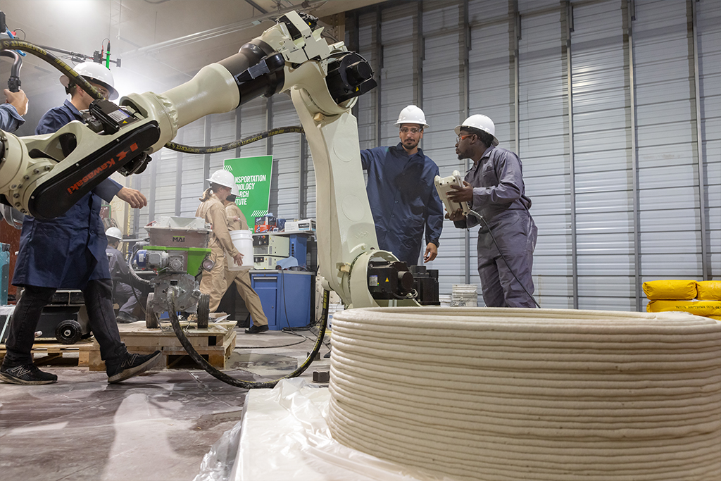 A group of researchers wearing hard hats and other protective gear work around a large robotic arm that is 3D printing with concrete.