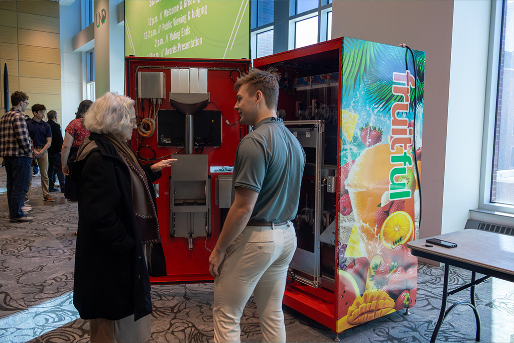 A student stands next to an open prototype of a smoothie vending machine at an expo.