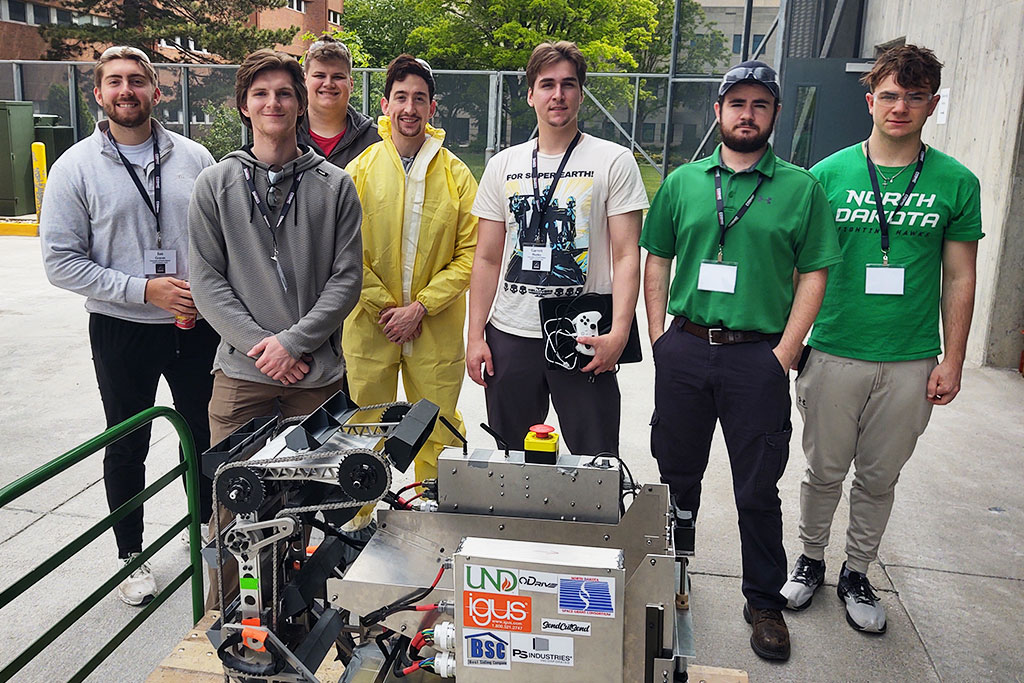 Seven students line up for a group photo with a robot in the foreground. They all smile, some wearing UND green shirts and some protective gear.