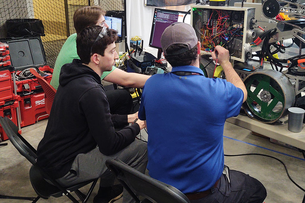 Three students work on a lunar robot, with the robot's wires and inner workings revealed.