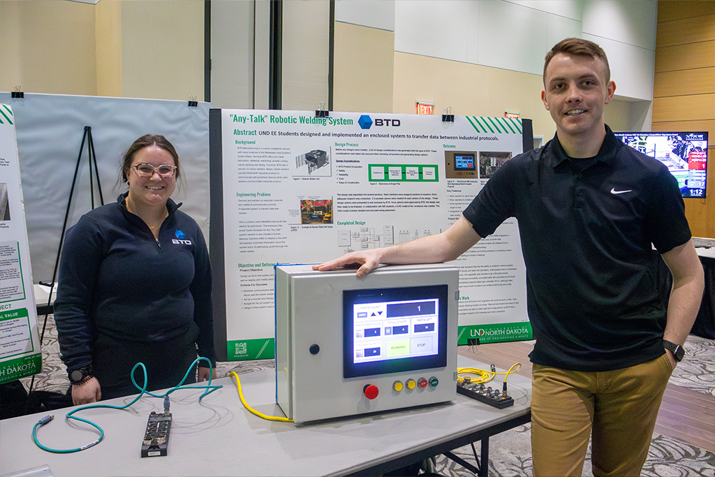 Two students smile in front of their engineering design poster titled "Any-Talk Robotic Welding System - BTD." The student on the right places their hand on a rectangular module with an LCD screen and buttons connected to other machine components.