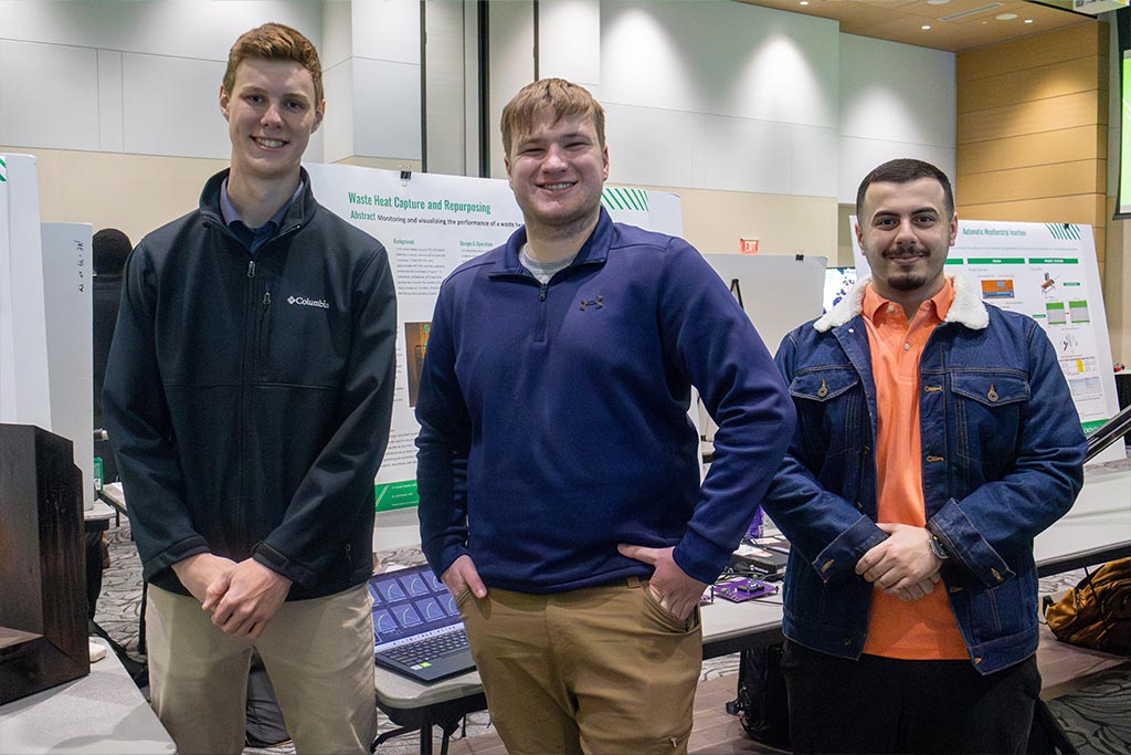 Three students smile for a group photo in front of an engineering design poster titled "Waste Heat Capture and Repurposing."