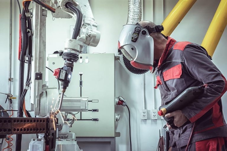 A person wears a welding mask as they supervise the autonomous operation of a robotics welder arm welding a piece of metal.