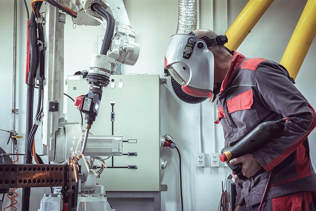 A person wears a welding mask as they supervise the autonomous operation of a robotics welder arm welding a piece of metal.