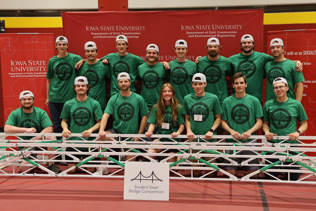 Fourteen UND students wearing kelly green shirts stand in front of a bridge structure. They smile and put their arms over each other's shoulders.