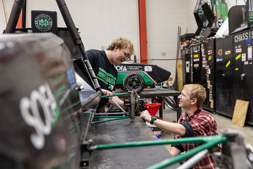 Two students smile and laugh as they work together in a high bay lab space dedicated to building race cars.