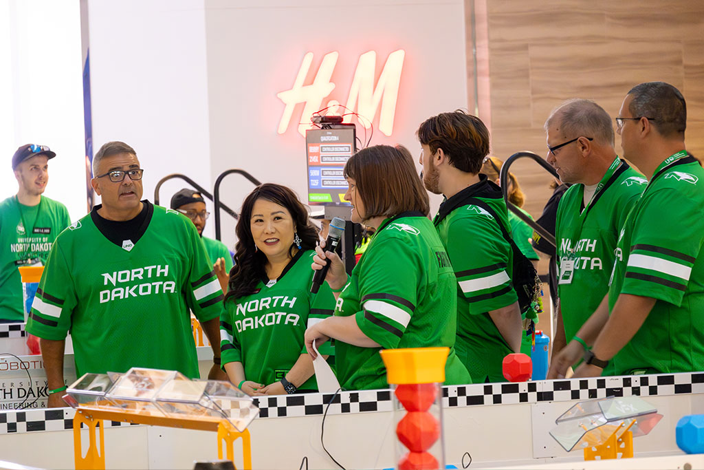 Judges for a robotics competition wear green UND hockey jerseys as they address the crowd during an opening ceremony.