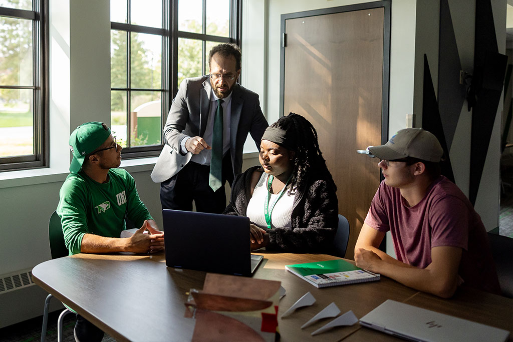 A professor talks with three students as they discuss something on a laptop inside of the Digital Engineering lab of the Mark & Claudia Thompson National Security Corridor.