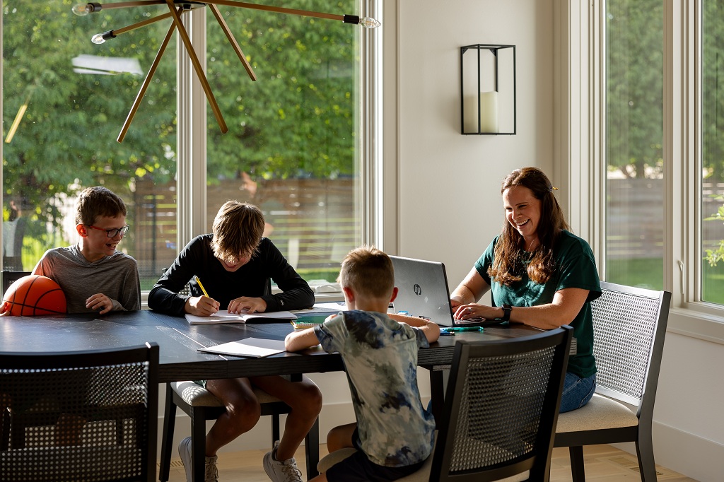 A family sits at a dining room table as a parent smiles and works at a laptop.