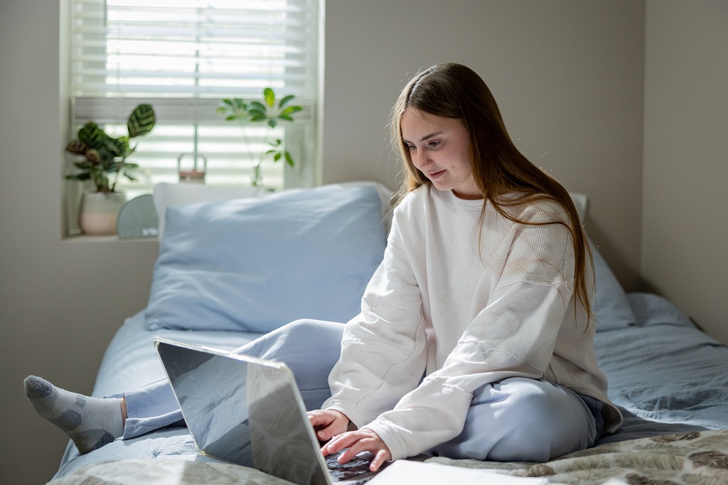 A young woman sits on a bed leisurely as she types on a computer.