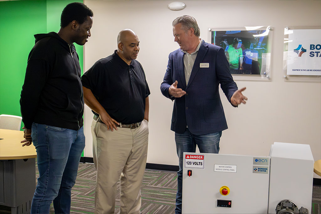 A man in a business suit talks to two other men, gesturing at equipment inside of a box.