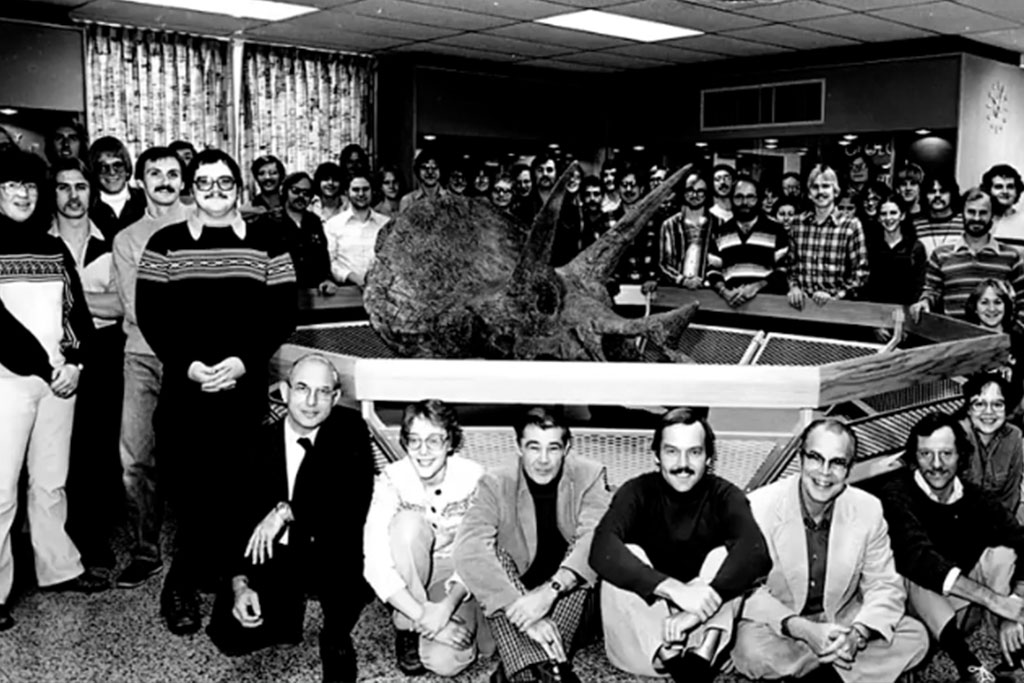 A black and white photograph of a large group of people surrounding a Triceratops skull in a museum.