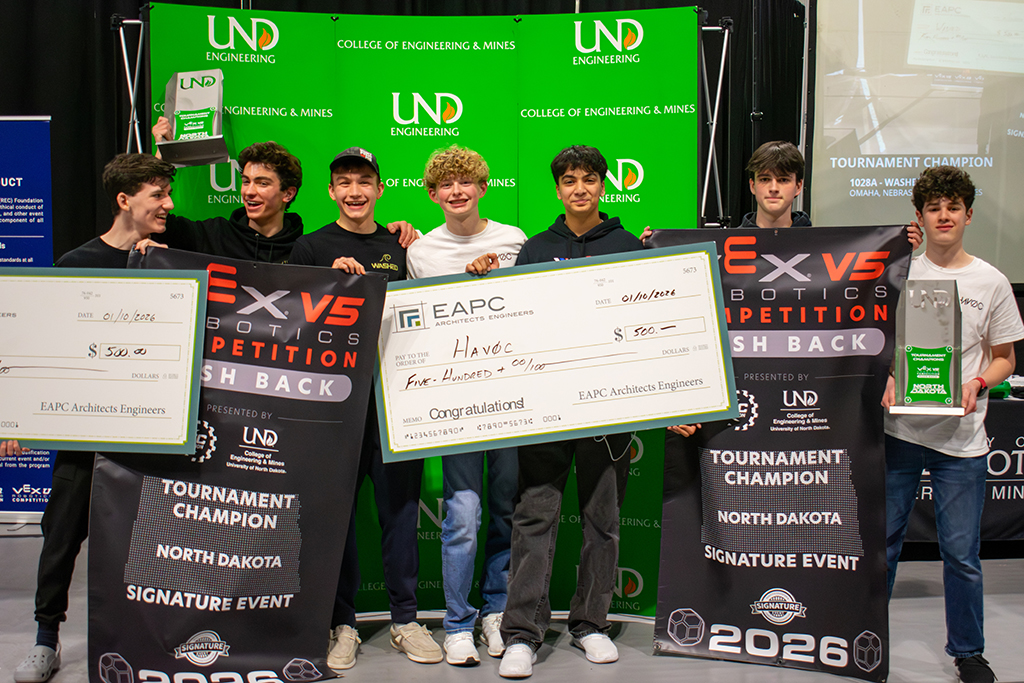 Seven high schoolers smile in front of a UND College of Engineering & Mines backgrop as they hold large checks presented by EAPC Architects Engineers, VEX V5 Robotics Competition Tournament Champion banners, and a large tournament champions trophy with the University of North Dakota logo.