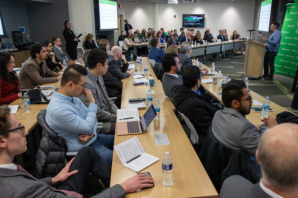 A classroom full of researchers and professors listen to a speaker at a podium in the front of the room.