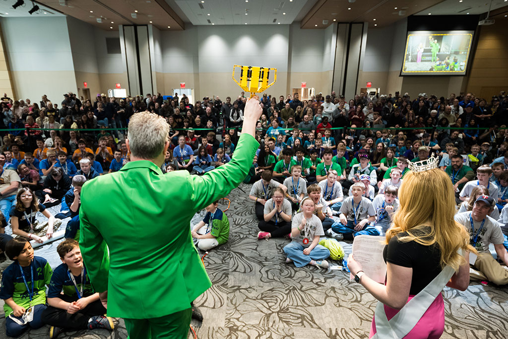 A man in a green suit jacket holds up a yellow trophy made of LEGO bricks to a room full of children.