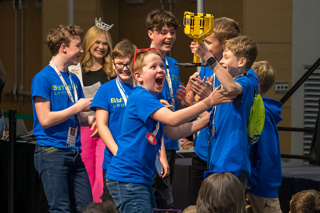 Children smile brightly as they hold up a yellow trophy made out of LEGO bricks.
