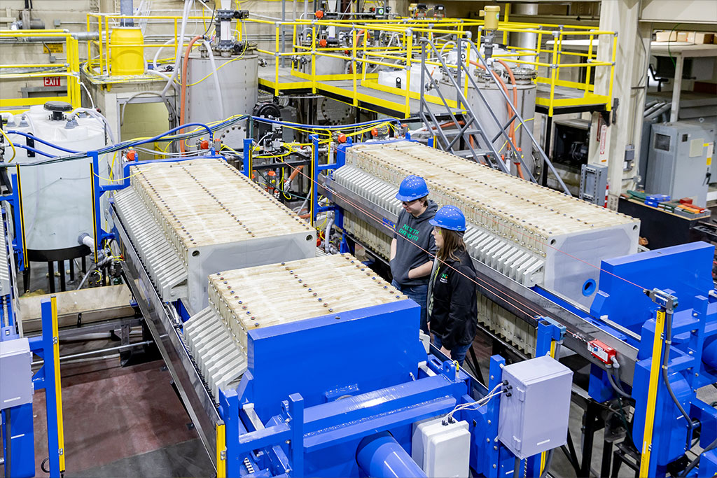 Two researchers stand in front of large laboratory equipment inside of a large industrial-scale research facility.
