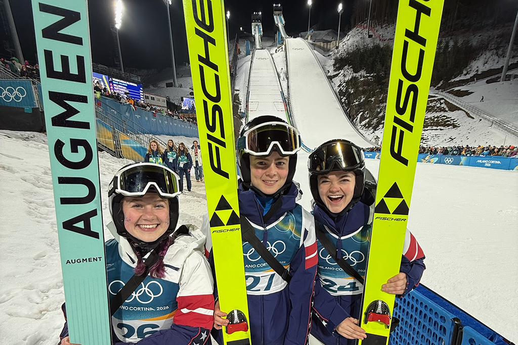 Three Olympic ski jumpers stand next to each other and smile for a photo in front of the ski jump.