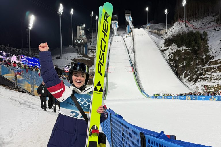 A ski jumper, Paige Jones, stands next to an Olympic ski jump with her fist raised in the air triumphantly.