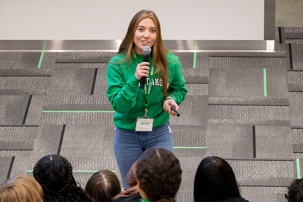 A University of North Dakota student in a green sweater smiles and she talks into a microphone in front of a group of children at the Memorial Union Social Staircase.