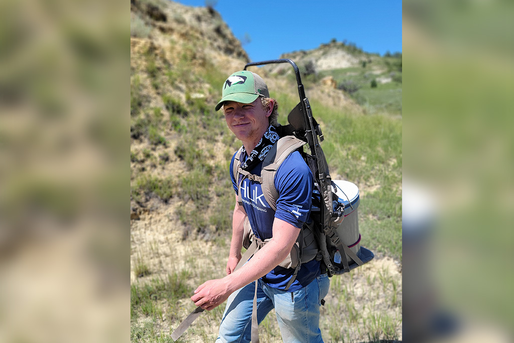 A student wearing a University of North Dakota Fighting Hawks baseball cap smiles as he walks across a remote North Dakota landscape with equipment strapped to his back.