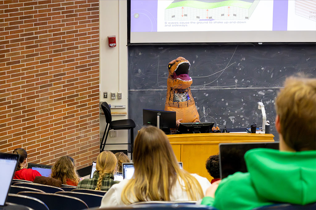 A person in a dinosaur costume instructs at a podium to students inside of a lecture hall.