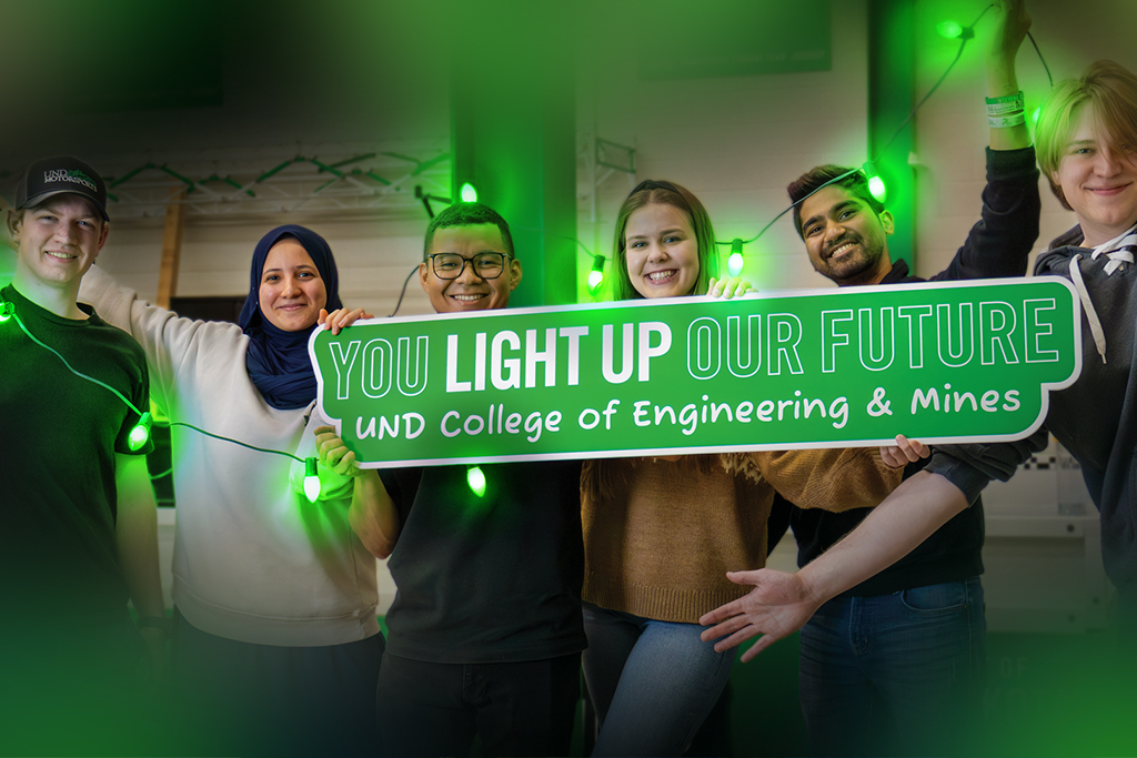 Students smile and hold up a sign that reads: "You light up our future | UND College of Engineering & Mines" while also holding up bright green string lights inside of a University of North Dakota laboratory.