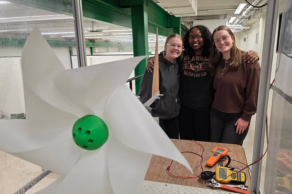 Three University of North Dakota students smile and hug eachother next to their project inside of a College of Engineering & Mines laboratory.