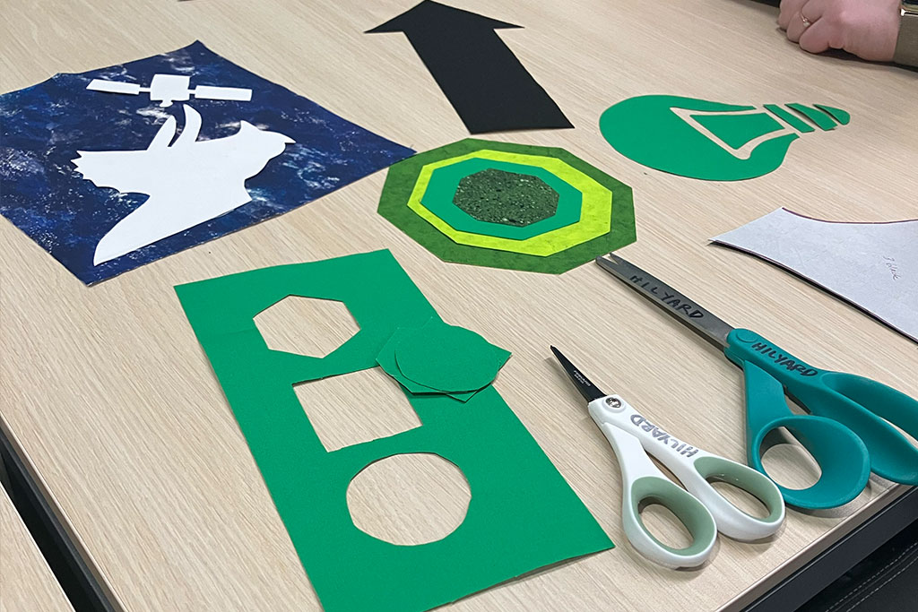 Pieces of cloth and scissors lay on a table. Some cut pieces of cloth are geometic while a few are distinctly a dinosaur, a satelitte, and an arrow.