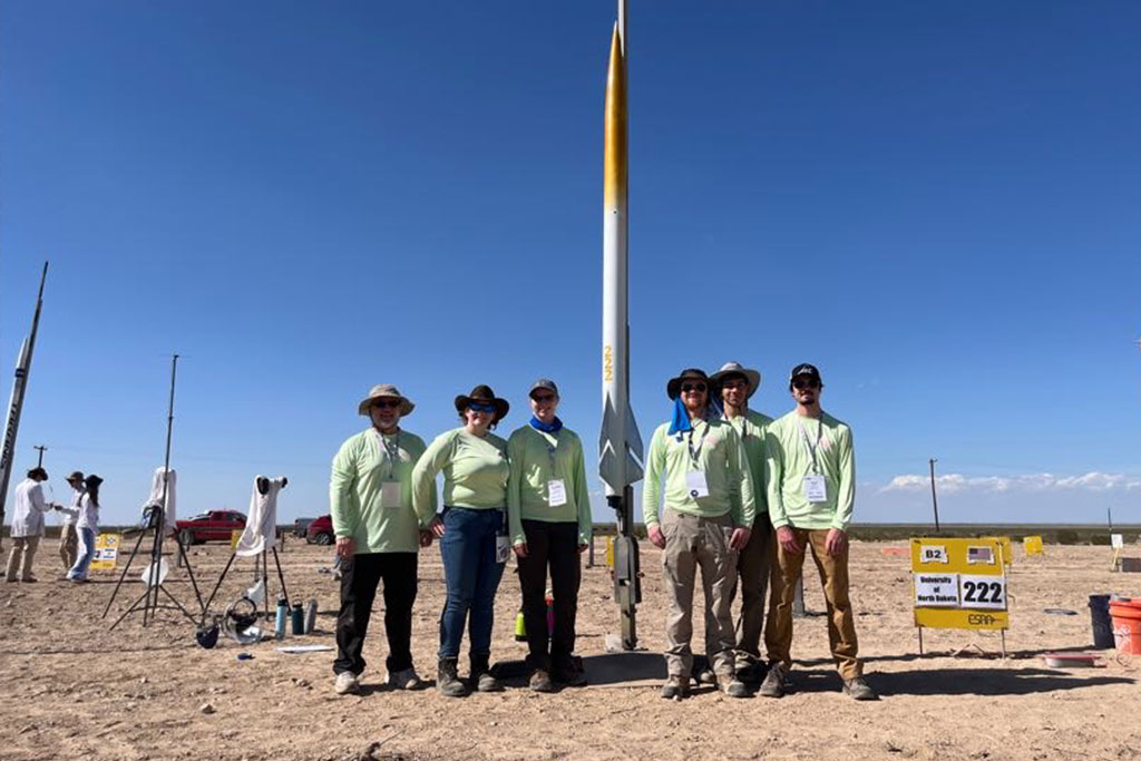 Six students on the University of North Dakota Advanced Rocketry team stand for a group photo in the desert next to their rocket that is prepped for launch in a national competition.