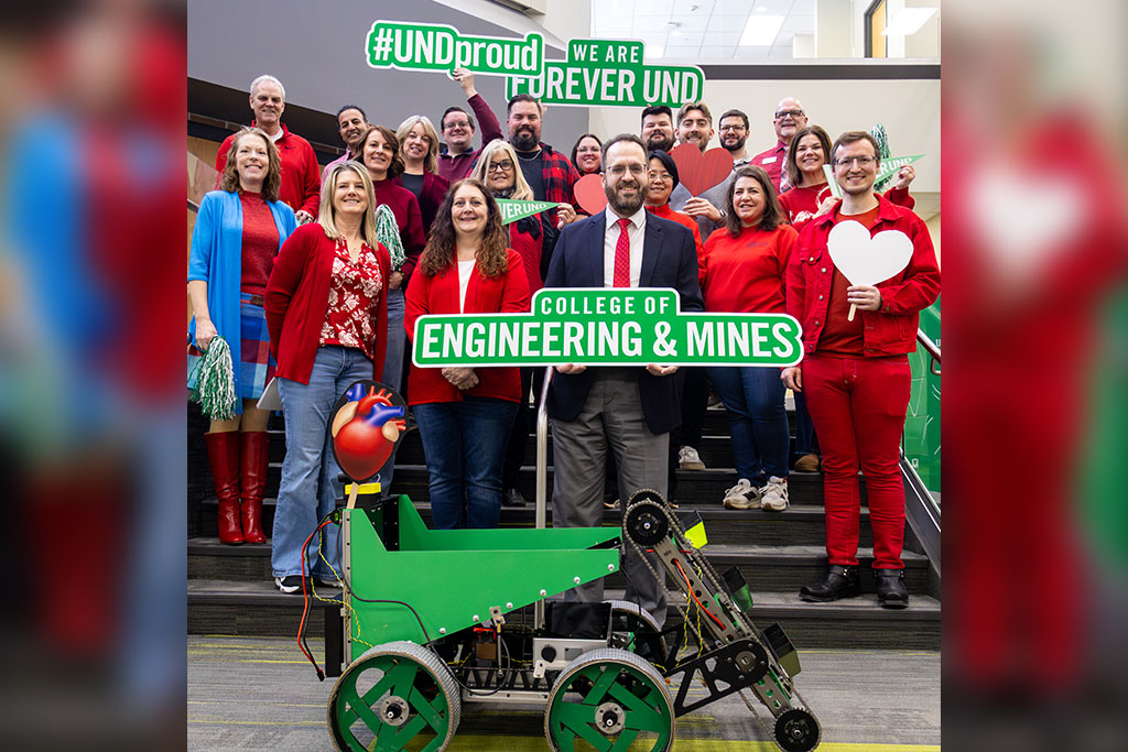 Faculty, staff, and students of the University of North Dakota College of Engineering & Mines wear red clothing for a group photo. In front of them in a lunar mining robot built by students, adorned with a decorative heart cut out. The people hold heart-shaped cut outs and signs that read: "College of Engineering & Mines," "#UNDproud," and "Forever UND."