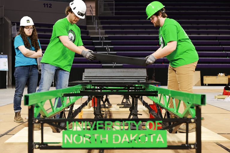Two students wearing UND green shirts and hard hats assemble a model steel bridge. Text on the bridge reads: "University of North Dakota."